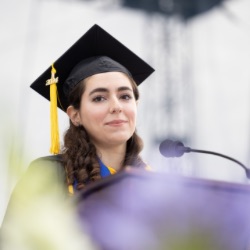 A young woman with long, dark hair wears a black graduation cap with a yellow tassel and gown. She stands at a podium with a microphone, appearing to give a speech. 的 background is blurred, emphasizing her as the focal point.